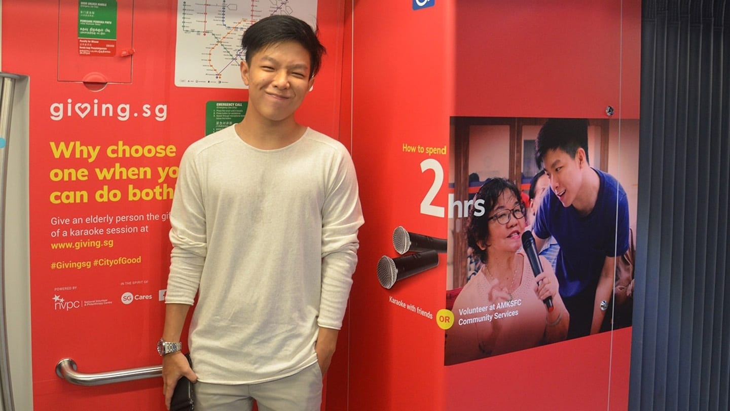 man in white shirt posing with a giving.sg ad on mrt