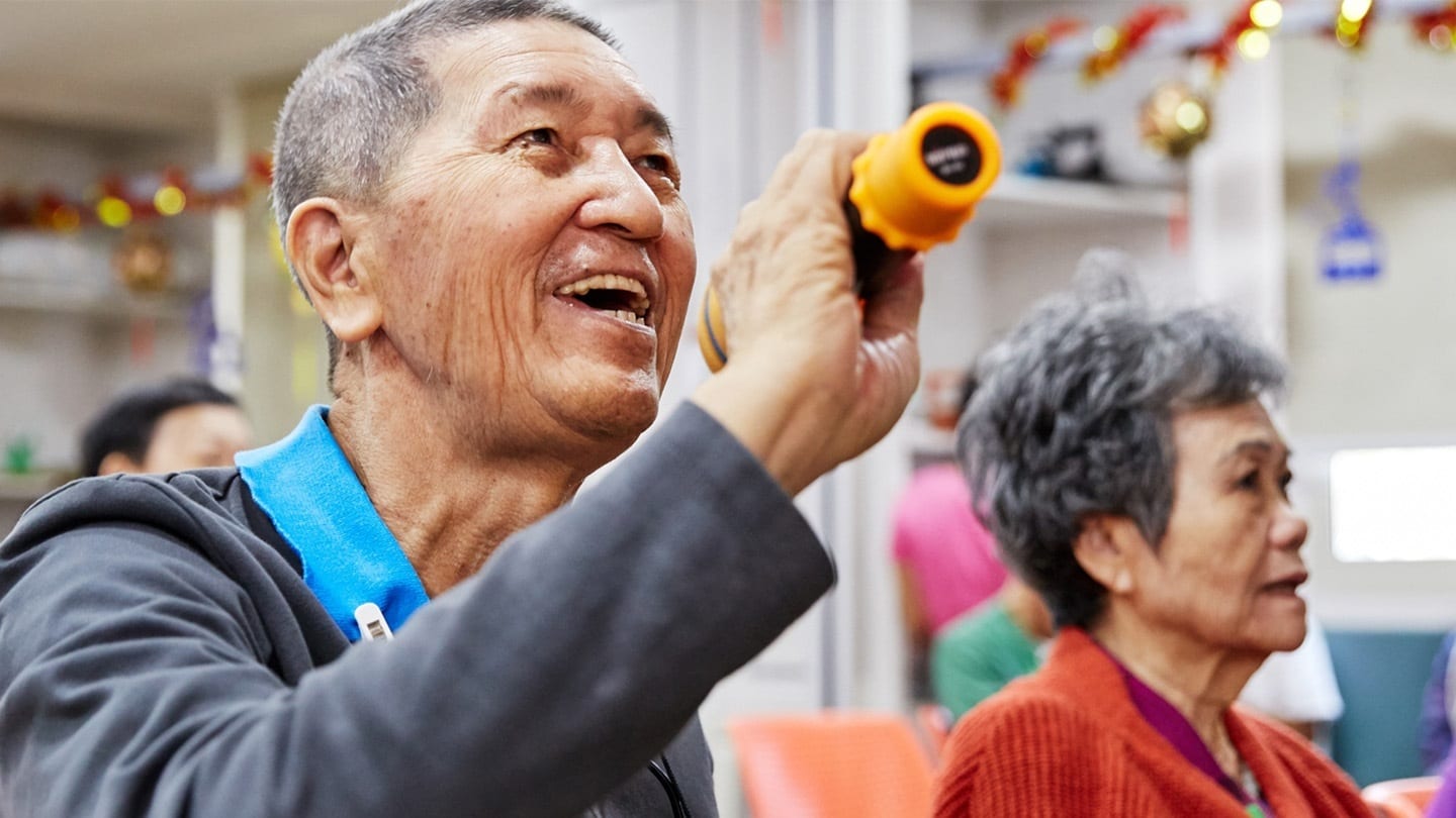 elderly man in long-sleeved grey shirt with blue collar singing into a microphone