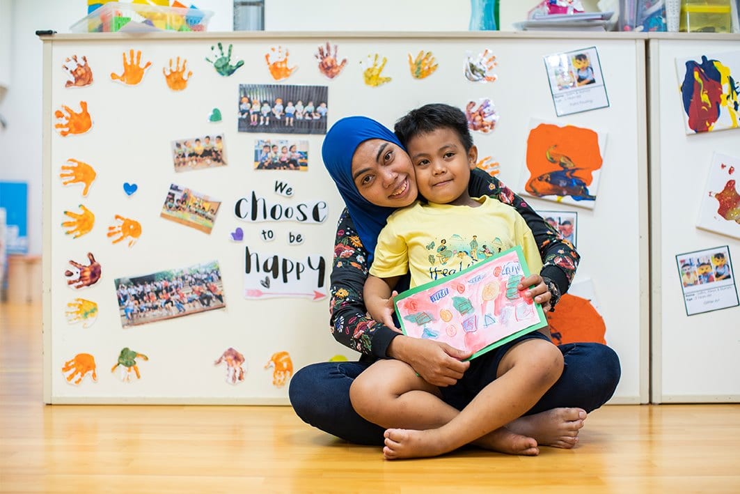 woman in blue tudung and boy in yellow shirt in front of wall saying we choose to be happy