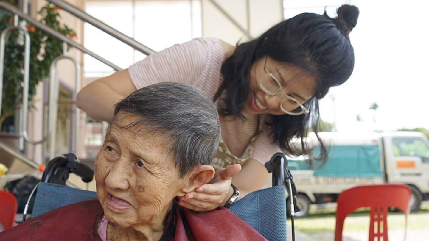 volunteer in transparent glasses and pink striped shirt touching hair of elderly lady in wheelchair