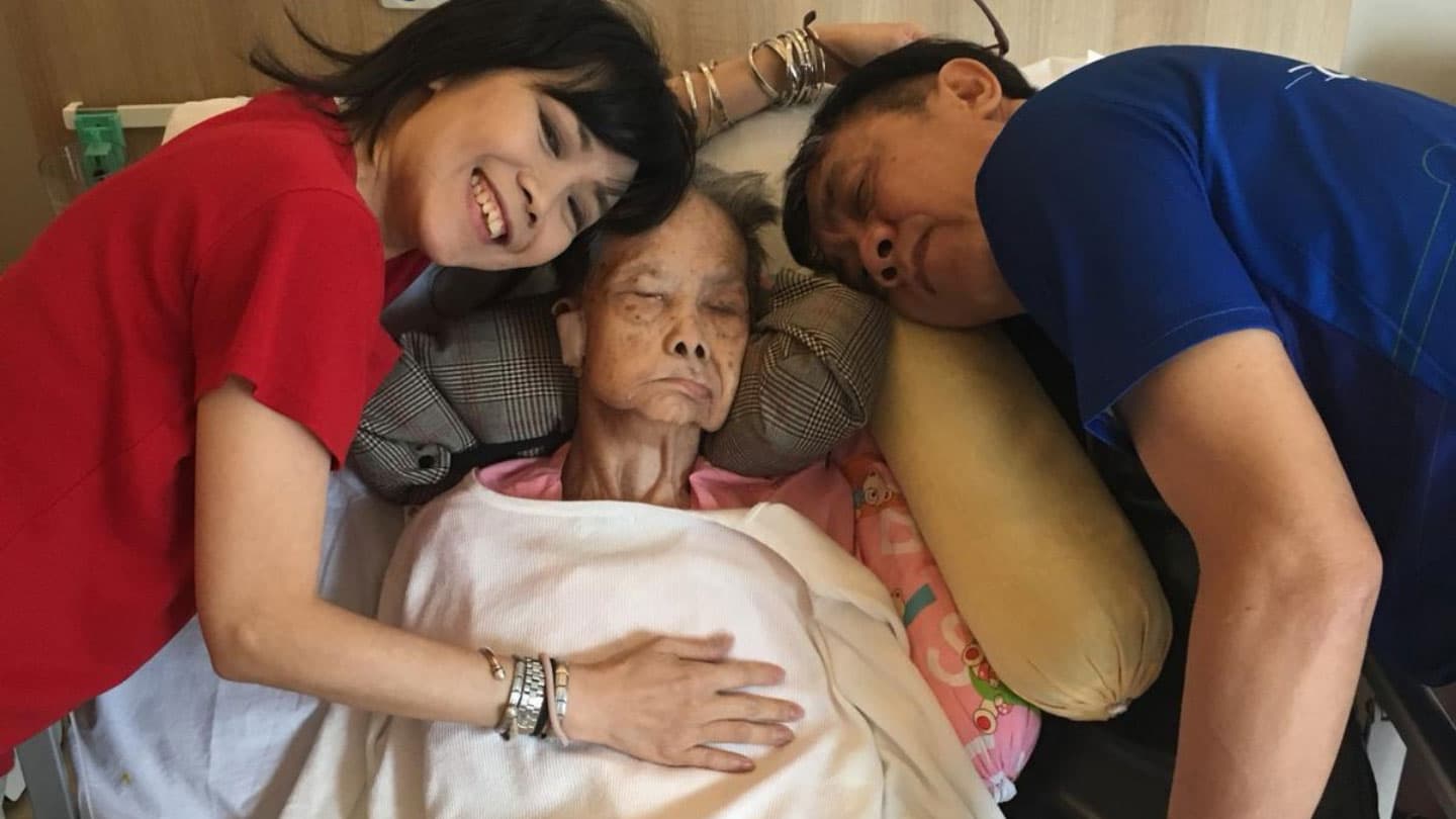 two female volunteers in red and blue shirts leaning on elderly women lying in hospice bed