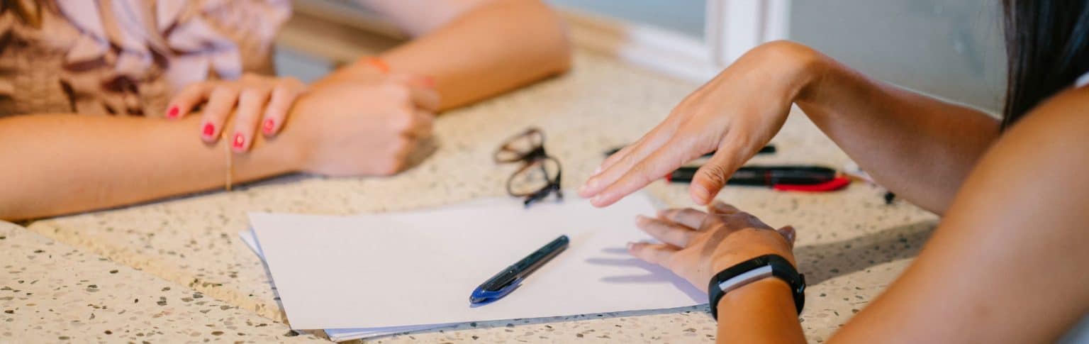 woman in short sleeves sitting opposite woman with red fingernails with papers between them