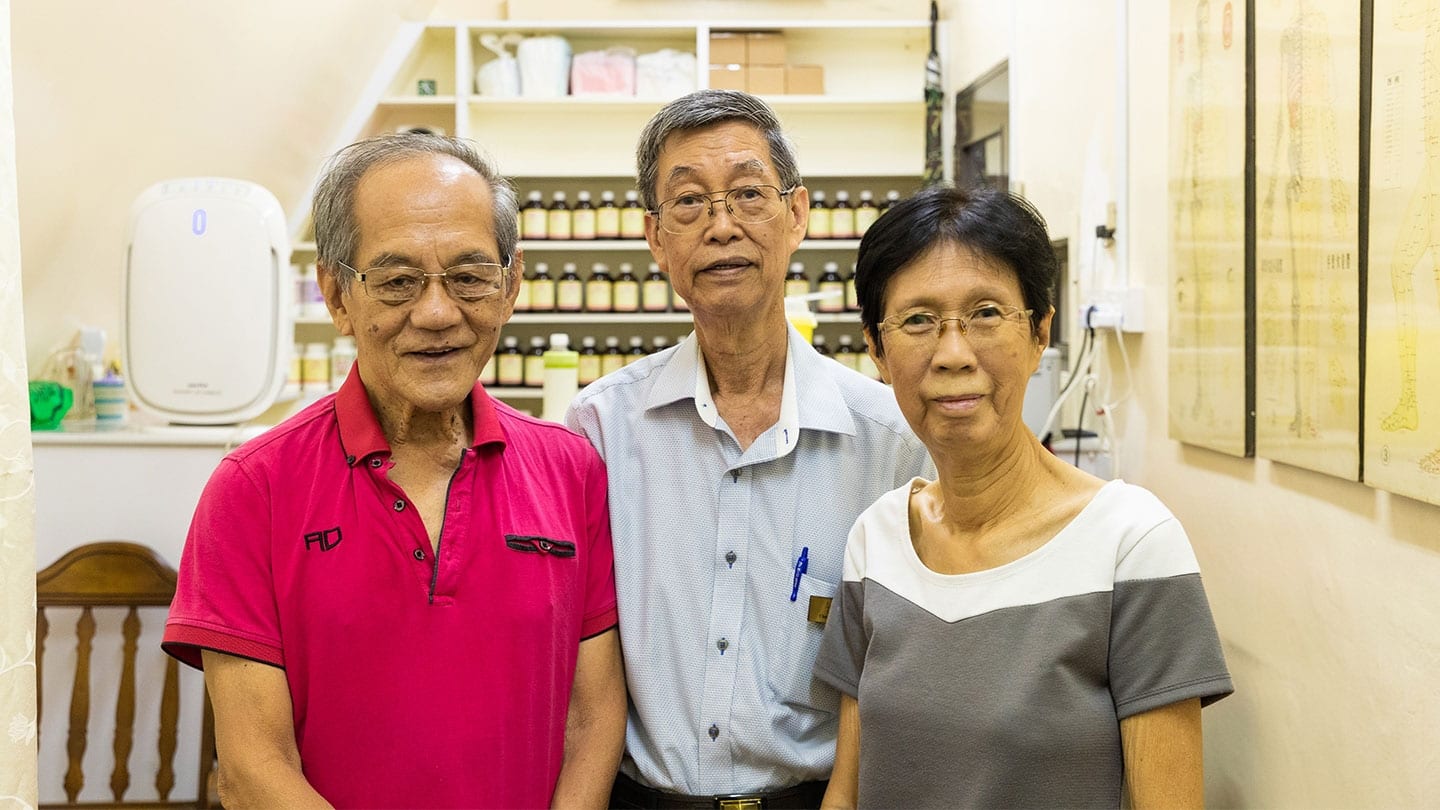 two elderly men and an elderly woman standing in front of a cabinet containing shelves of bottles