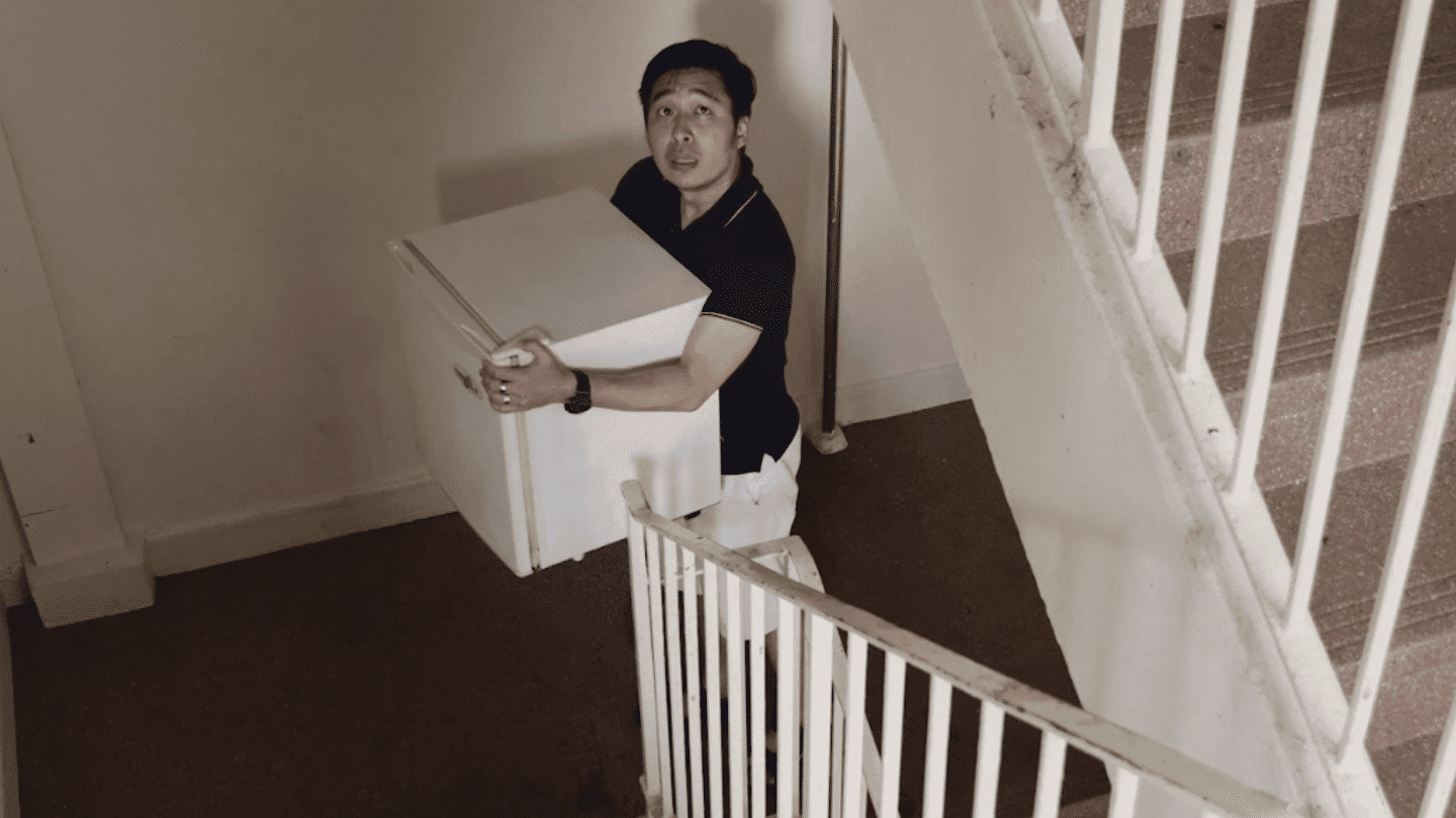 black and white photo featuring a man in a black shirt carrying a mini-fridge up the stairs