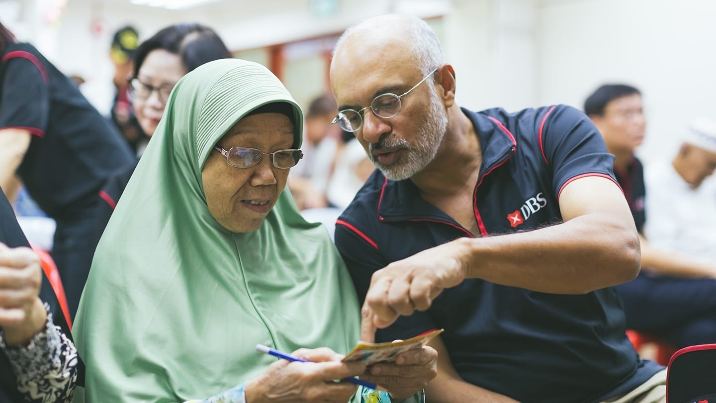 volunteer in dbs shirt chatting with elderly woman in green tudung