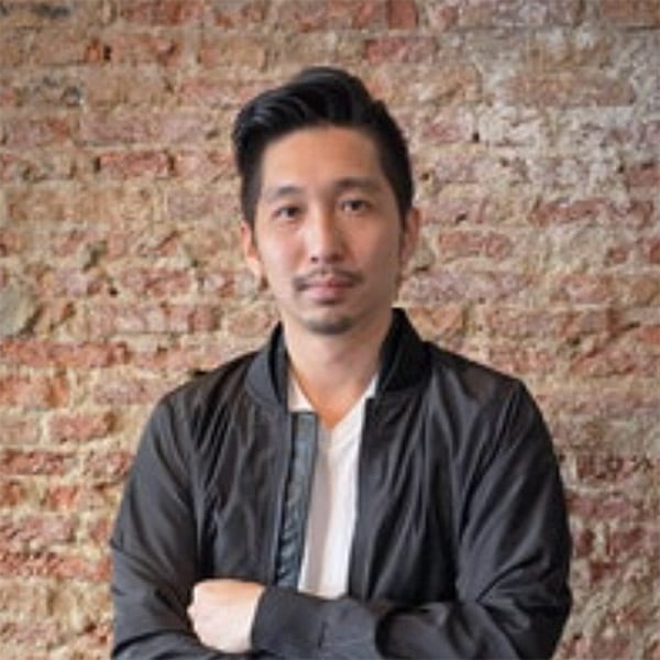 moustached young man in black jacket standing in front of a red brick wall