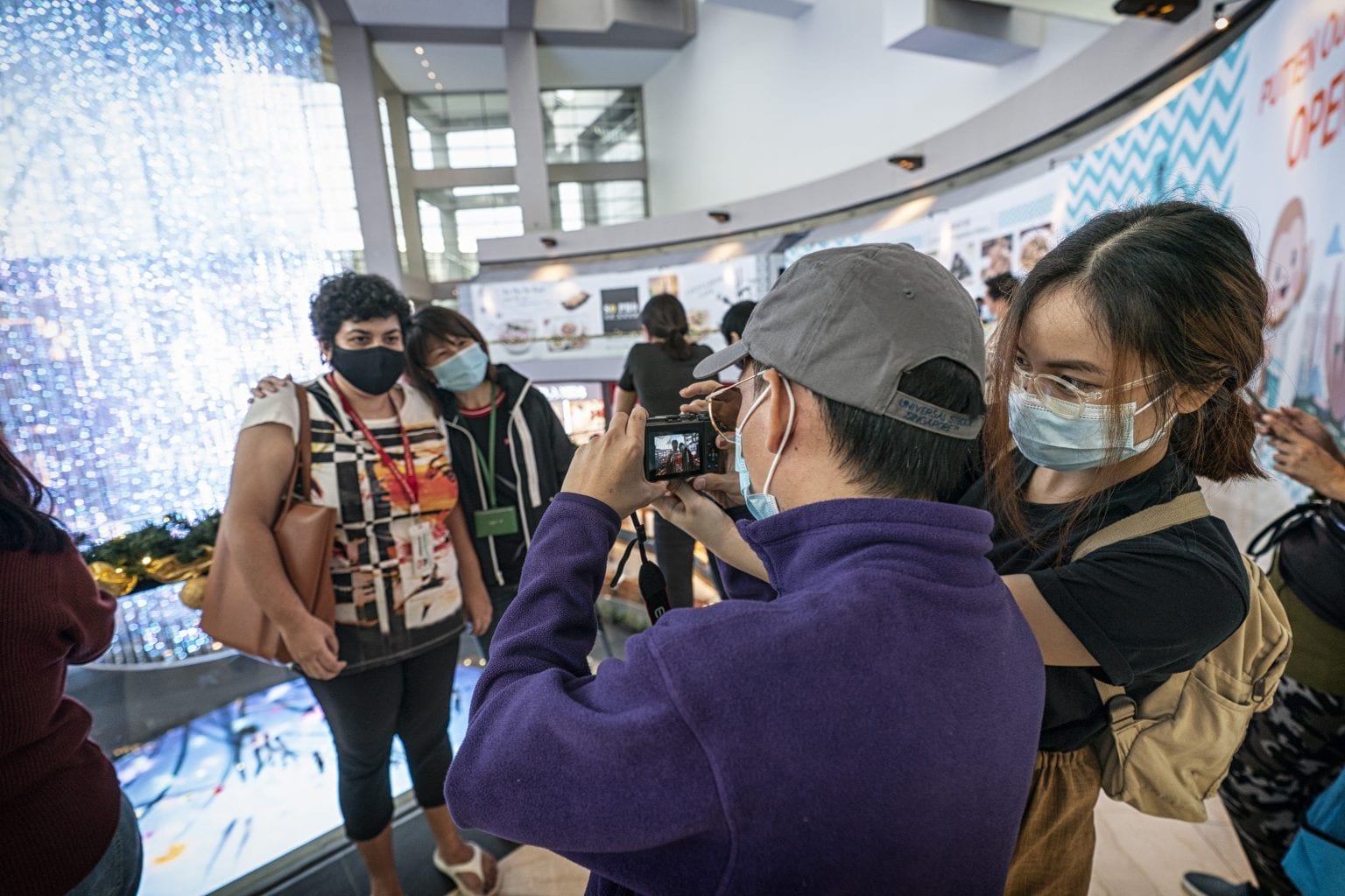 man in grey cap and girl with backpack helping two ladies take a photo in front of a glittering exhibition