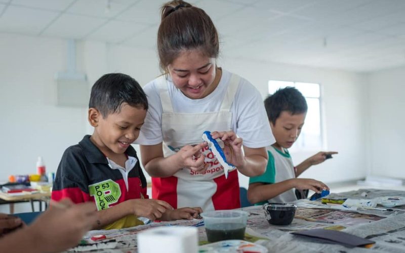 young female volunteer in red and white apron doing arts and crafts with two boys