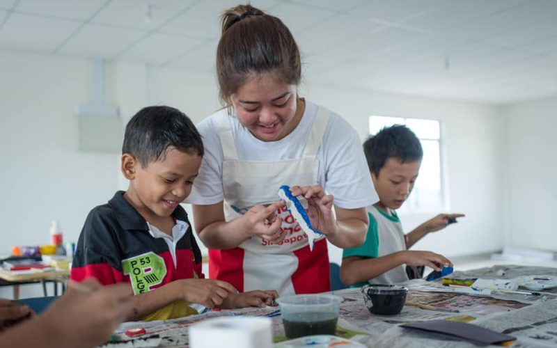 young female volunteer in red and white apron doing arts and crafts with two boys